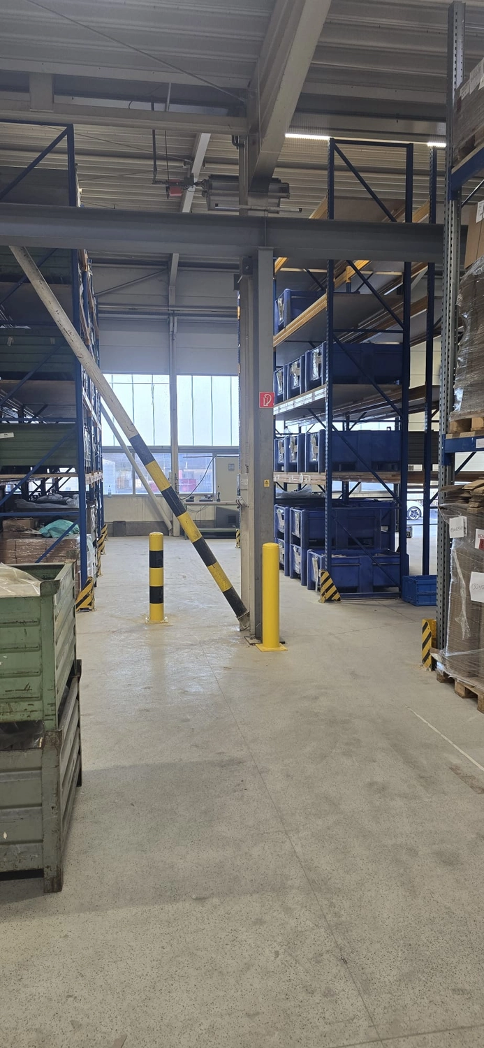 MTHH GbR - Kabeltragsystembau, Regalbau/Prüfung und Tiefbau - Innenansicht einer Lagerhalle mit hochverdichteten Palettenregalen und Sicherheitsausrüstung. Im Vordergrund befindet sich ein großer Stützpfeiler mit gelben Sicherheitspollern an seinem Fuß und einem schwarz-gelb gestreiften diagonalen Träger. 