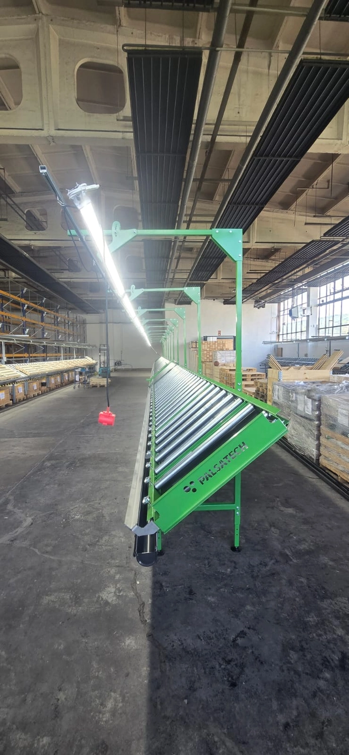 MTHH GbR - Kabeltragsystembau, Regalbau/Prüfung und Tiefbau - Eine helle, geräumige Lagerhalle mit hohen Decken und großen Fenstern auf der rechten Seite, durch die man Bäume und einen Hügel sieht. Zwei lange Reihen grün umrahmter Rollenbahnen erstrecken sich in die Ferne; am Ende der linken Reihe steht eine Person. Links befinden sich hohe Industrieregale, rechts Holzpaletten und mit Plastikfolie umwickelte Kisten. Sonnenlicht strömt durch die Fenster und wirft lange Schatten auf den Betonboden.