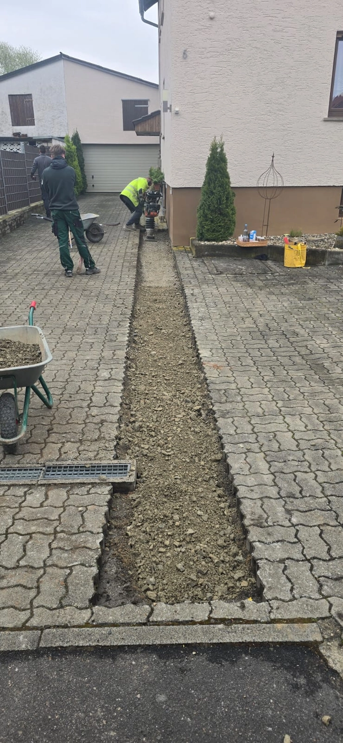 MTHH GbR - Kabeltragsystembau, Regalbau/Prüfung und Tiefbau - Bauarbeiter entfernen einen schmalen Streifen aus Verbundpflastersteinen von einer Wohnhauseinfahrt, um einen Graben auszuheben, wahrscheinlich für die Verlegung oder Reparatur von Versorgungsleitungen.