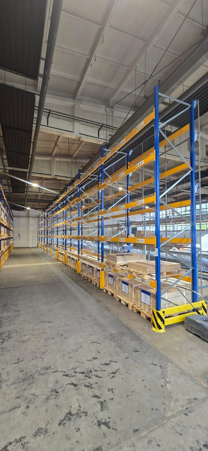 MTHH GbR - Kabeltragsystembau, Regalbau/Prüfung und Tiefbau - In einer geräumigen Lagerhalle mit Betonboden und hohen Decken steht eine lange Reihe blauer und gelber Industriepalettenregale.