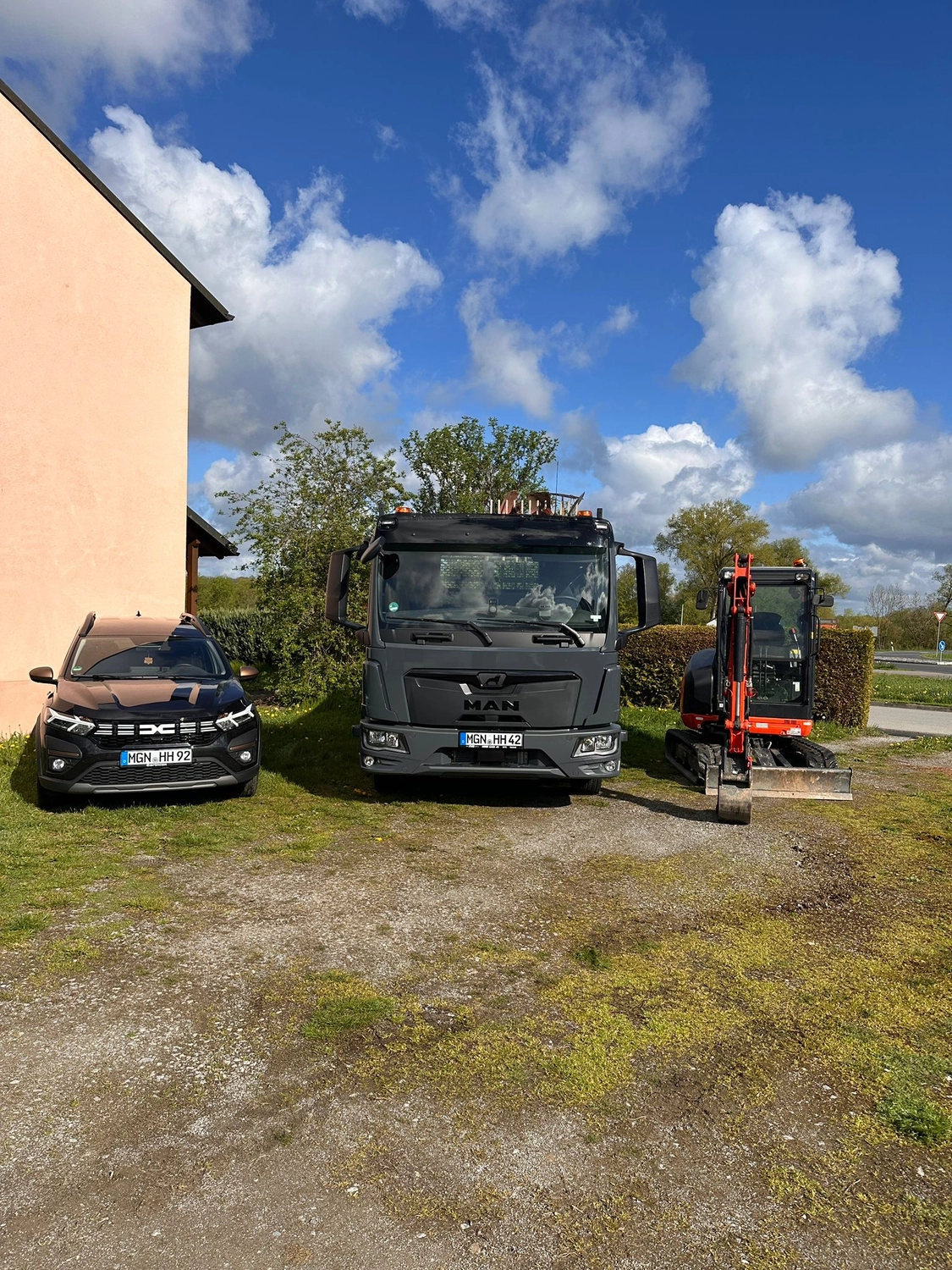 MTHH GbR - Kabeltragsystembau, Regalbau/Prüfung und Tiefbau - Ein schwarzer Pkw, ein großer grauer Lastwagen und ein orangefarbener Minibagger stehen nebeneinander auf einem Schotter- und Grasplatz vor einem rosafarbenen Gebäude unter einem blauen Himmel mit flauschigen weißen Wolken.