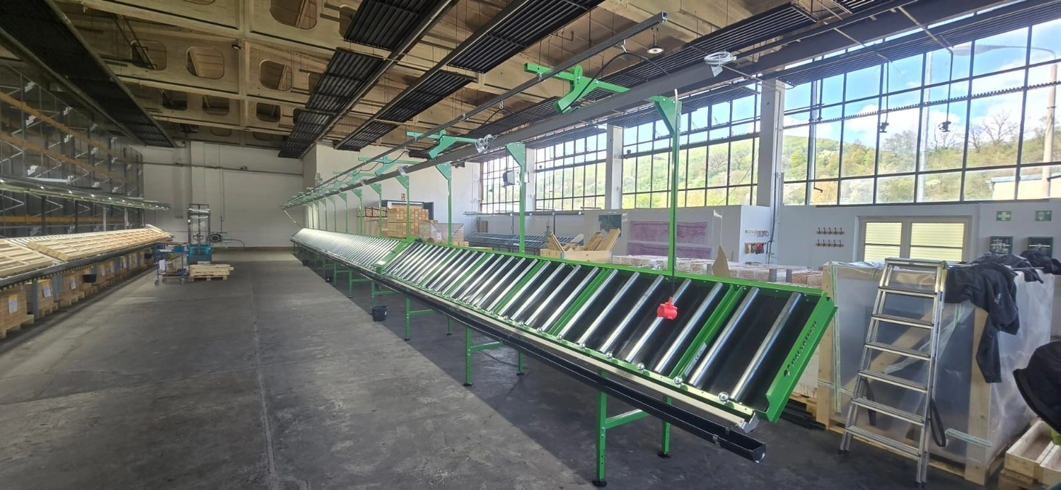 MTHH GbR - Kabeltragsystembau, Regalbau/Prüfung und Tiefbau - Eine helle, geräumige Lagerhalle mit hohen Decken und großen Fenstern auf der rechten Seite, durch die man Bäume und einen Hügel sieht. Zwei lange Reihen grün umrahmter Rollenbahnen erstrecken sich in die Ferne; am Ende der linken Reihe steht eine Person. Links befinden sich hohe Industrieregale, rechts Holzpaletten und mit Plastikfolie umwickelte Kisten. Sonnenlicht strömt durch die Fenster und wirft lange Schatten auf den Betonboden.