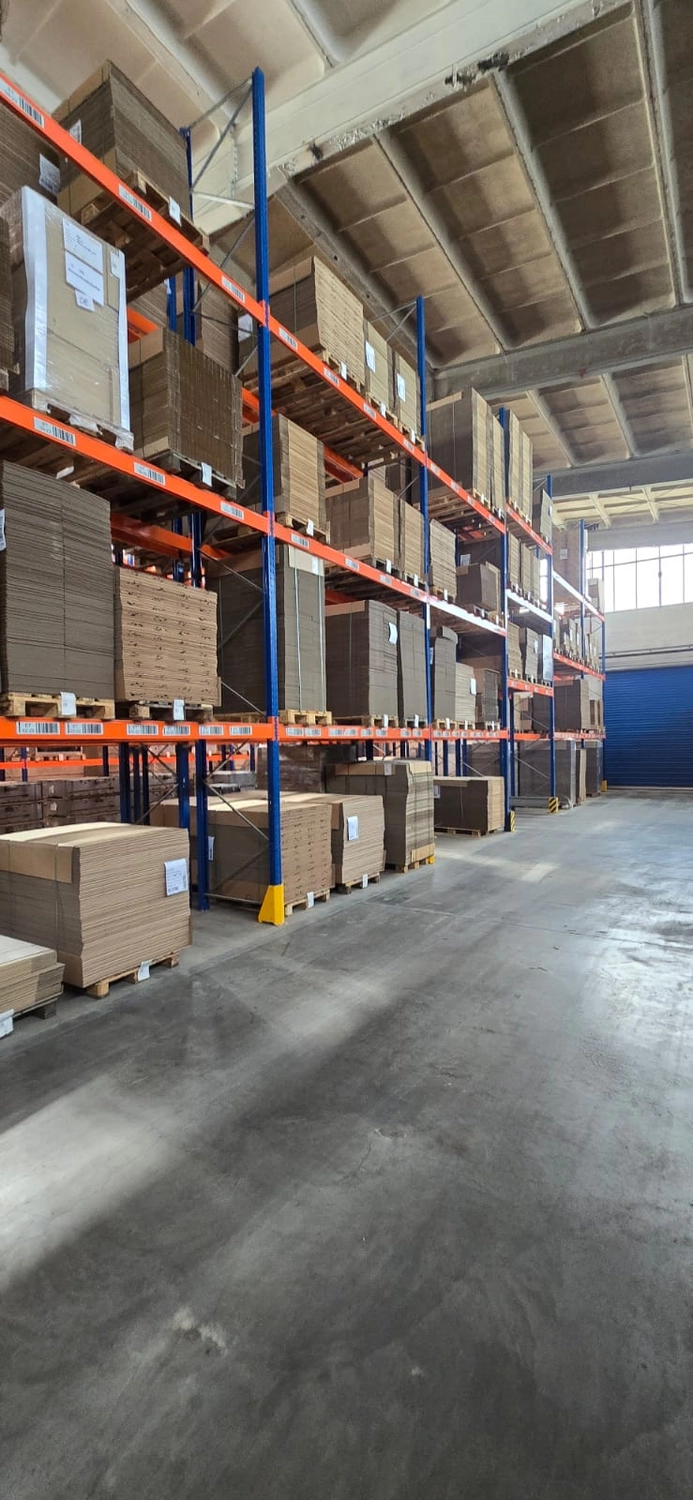 MTHH GbR - Kabeltragsystembau, Regalbau/Prüfung und Tiefbau - Eine Weitwinkelaufnahme einer Lagerhalle mit hohen Decken, auf der hohe Industrieregale mit Kartonbögen und -kisten stehen; im Vordergrund ist ein polierter Betonboden zu sehen.
