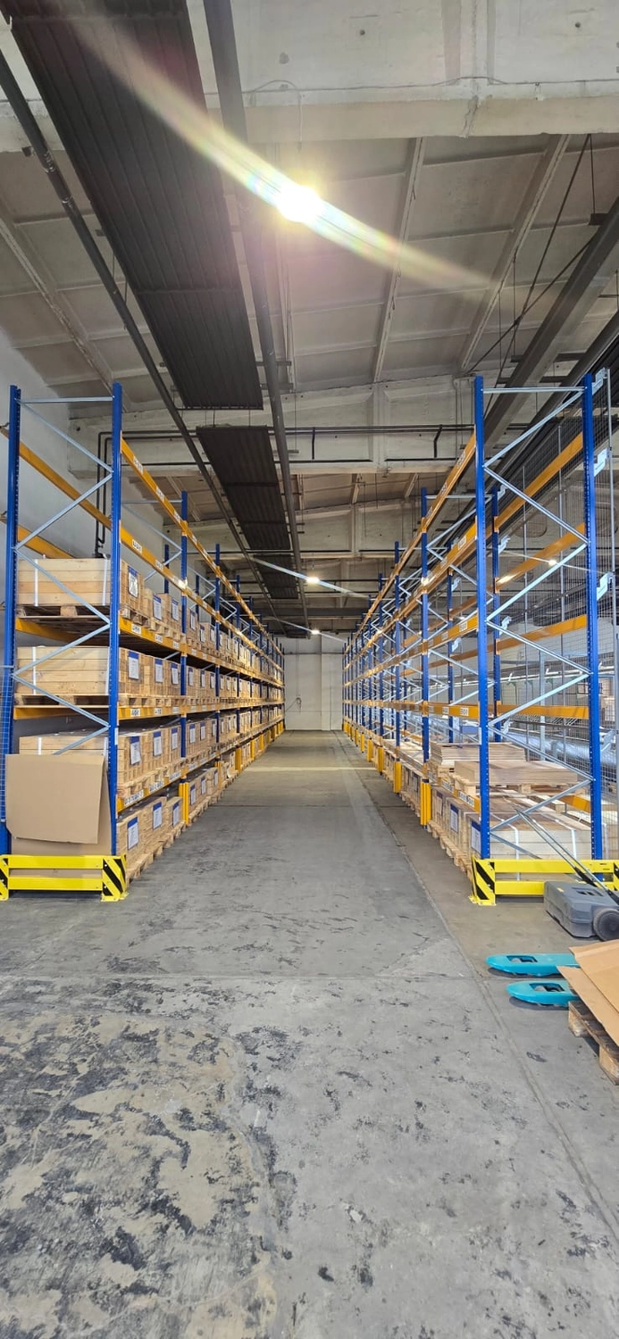 MTHH GbR - Kabeltragsystembau, Regalbau/Prüfung und Tiefbau - Das Innere einer Lagerhalle zeigt zwei lange Reihen blauer und gelber Industrieregale, die mit Holzkisten gefüllt sind und durch einen breiten Betongang getrennt sind.