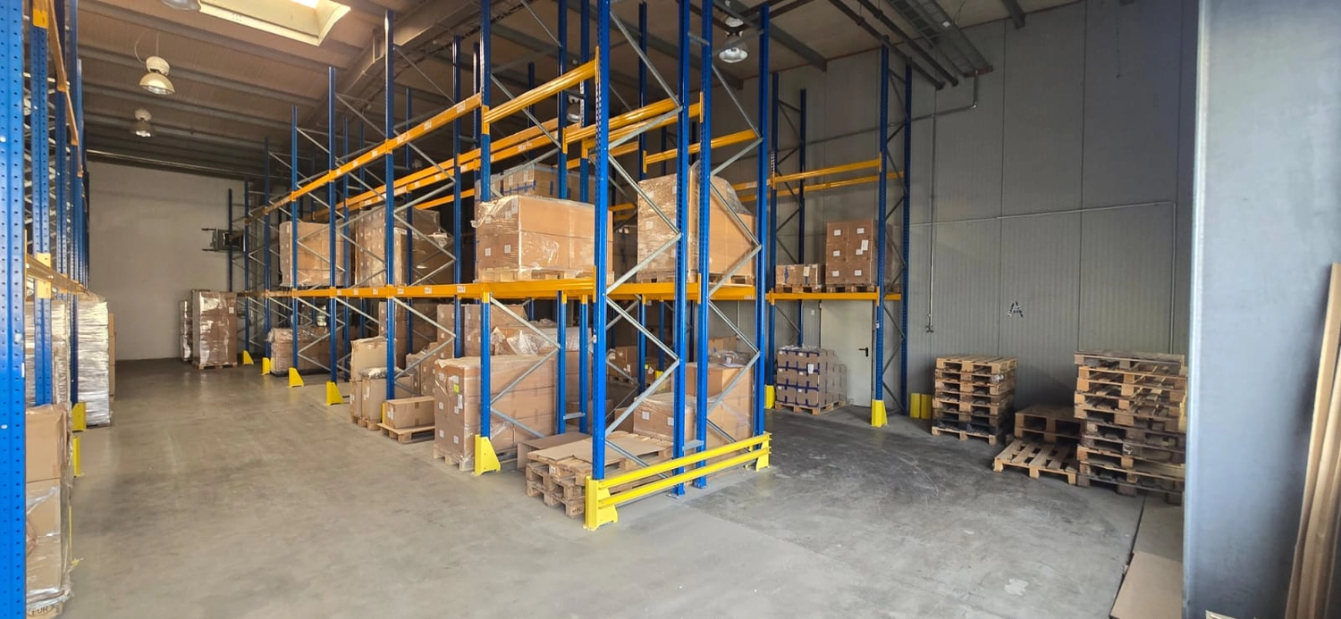 MTHH GbR - Kabeltragsystembau, Regalbau/Prüfung und Tiefbau - Diese Weitwinkelaufnahme zeigt das Innere einer Lagerhalle mit hohen, blau-gelben Industrieregalen, die mit verschiedenen Kartons und verpackten Waren gefüllt sind. Auf dem Betonboden vor einer grauen Wand sind außerdem Stapel von Holzpaletten zu sehen.