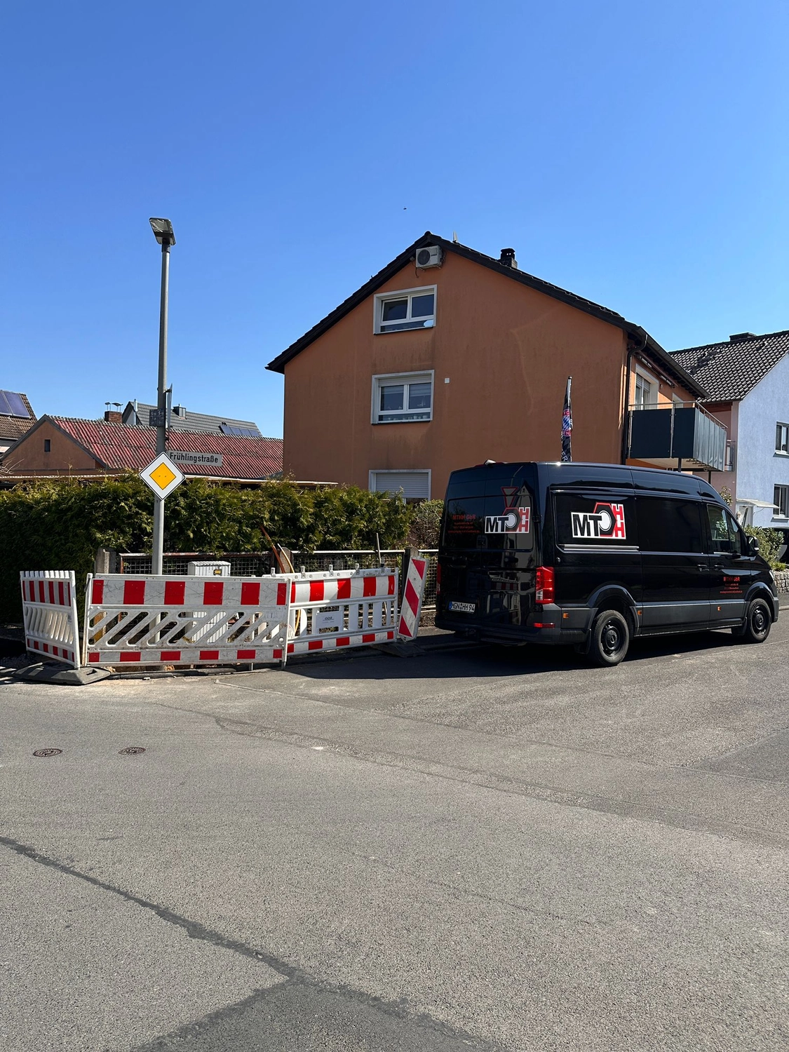 MTHH GbR - Kabeltragsystembau, Regalbau/Prüfung und Tiefbau - Eine vertikale Aufnahme auf Augenhöhe zeigt einen schwarzen Lieferwagen, der an einem sonnigen Tag in Deutschland auf einer asphaltierten Straße neben einer rot-weißen Baustellenabsperrung parkt. Hinter der Absperrung befindet sich eine grüne Hecke, und im Hintergrund erhebt sich unter strahlend blauem Himmel ein pfirsichfarbenes dreistöckiges Gebäude.