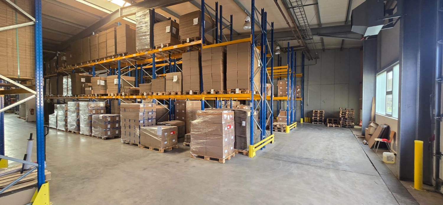 MTHH GbR - Kabeltragsystembau, Regalbau/Prüfung und Tiefbau - Das Innere einer Lagerhalle zeigt hohe, blau und gelb lackierte Metallregale, die mit zahlreichen Kartons beladen sind, von denen einige auf Paletten auf dem Betonboden eingeschweißt sind.