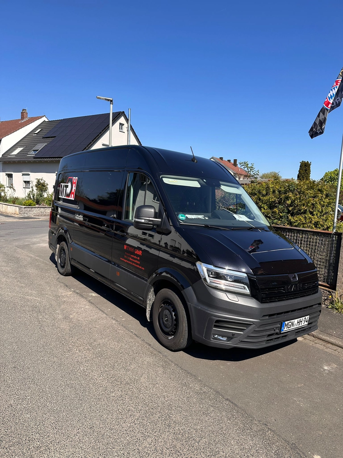 MTHH GbR - Kabeltragsystembau, Regalbau/Prüfung und Tiefbau - Ein schwarzer MAN TGE Kastenwagen parkt in einer Wohnstraße unter strahlend blauem Himmel. Der Wagen trägt rot-weiße Firmenaufdrucke an der Seite und ein deutsches Kennzeichen.