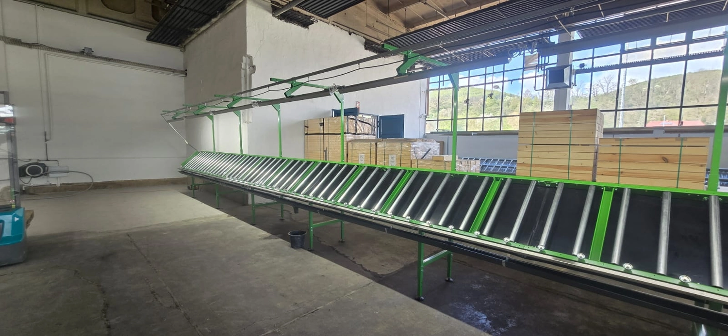 MTHH GbR - Kabeltragsystembau, Regalbau/Prüfung und Tiefbau - Eine helle, geräumige Lagerhalle mit hohen Decken und großen Fenstern auf der rechten Seite, durch die man Bäume und einen Hügel sieht. Zwei lange Reihen grün umrahmter Rollenbahnen erstrecken sich in die Ferne; am Ende der linken Reihe steht eine Person. Links befinden sich hohe Industrieregale, rechts Holzpaletten und mit Plastikfolie umwickelte Kisten. Sonnenlicht strömt durch die Fenster und wirft lange Schatten auf den Betonboden.