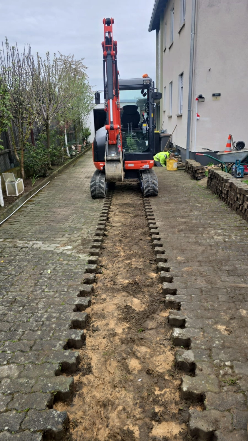 MTHH GbR - Kabeltragsystembau, Regalbau/Prüfung und Tiefbau - Am Ende eines langen, schmalen Grabens, der durch eine gepflasterte Auffahrt neben einem Haus gegraben wurde, steht ein orange-schwarzer Minibagger.