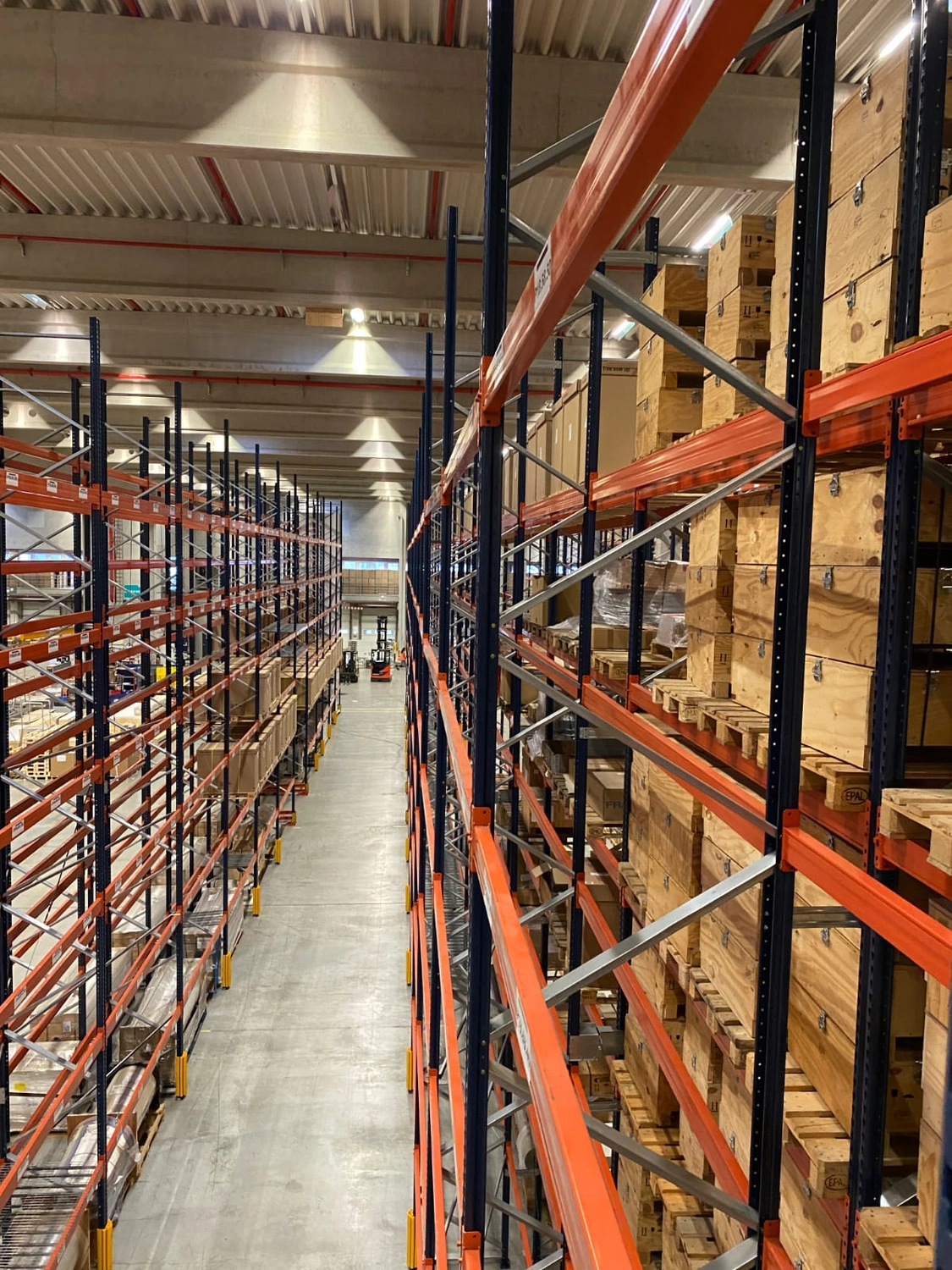 MTHH GbR - Kabeltragsystembau, Regalbau/Prüfung und Tiefbau - Eine Aufnahme aus der Vogelperspektive auf Augenhöhe zeigt einen Lagerhallengang, der mit hohen, orangefarbenen und blauen Industrieregalen aus Paletten gesäumt ist, die mit Holzkisten und -kartons gefüllt sind.