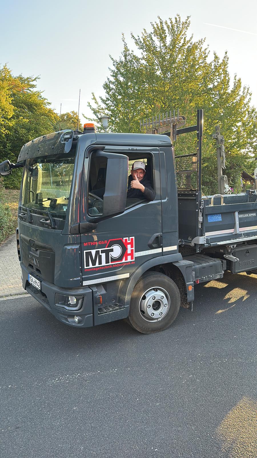 MTHH GbR - Kabeltragsystembau, Regalbau/Prüfung und Tiefbau - Ein Mann zeigt vom Fahrersitz eines dunkelgrauen MAN-Lkw mit dem Firmenlogo „MT H“ auf der Tür aus den Daumen nach oben.