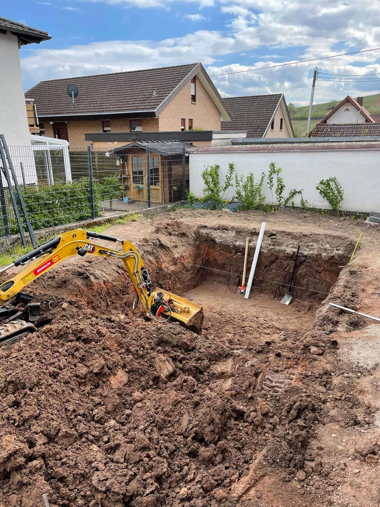 MTHH GbR - Kabeltragsystembau, Regalbau/Prüfung und Tiefbau - Ein Bagger hebt in einem Wohngarten, umgeben von Haufen frischer Erde und nahegelegenen Häusern, eine tiefe, rechteckige Grube aus.