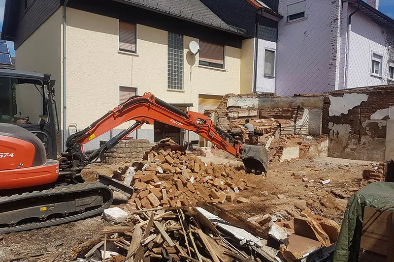 MTHH GbR - Kabeltragsystembau, Regalbau/Prüfung und Tiefbau - Ein orangefarbener Kubota-Bagger steht auf einer Baustelle, umgeben von Schutt- und Trümmerhaufen während des Abrisses eines Backsteingebäudes.