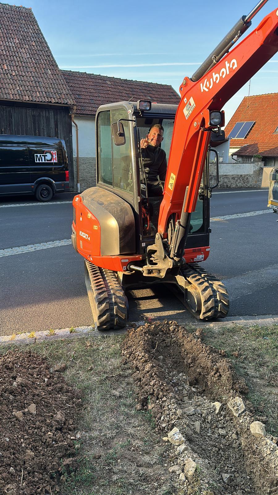 MTHH GbR - Kabeltragsystembau, Regalbau/Prüfung und Tiefbau - Ein Mann in der Fahrerkabine eines roten Kubota KX027-4 Minibaggers, der auf einer Straße neben einem frisch ausgehobenen Graben in einer Grasfläche geparkt ist.