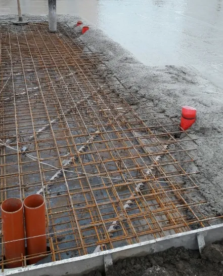 MTHH GbR - Kabeltragsystembau, Regalbau/Prüfung und Tiefbau - Eine Baustelle, auf der eine Stahlbewehrung für eine Betonplatte ausgelegt ist; im Hintergrund ragen orangefarbene PVC-Rohre aus dem Boden, und frischer Beton ist teilweise eingegossen.