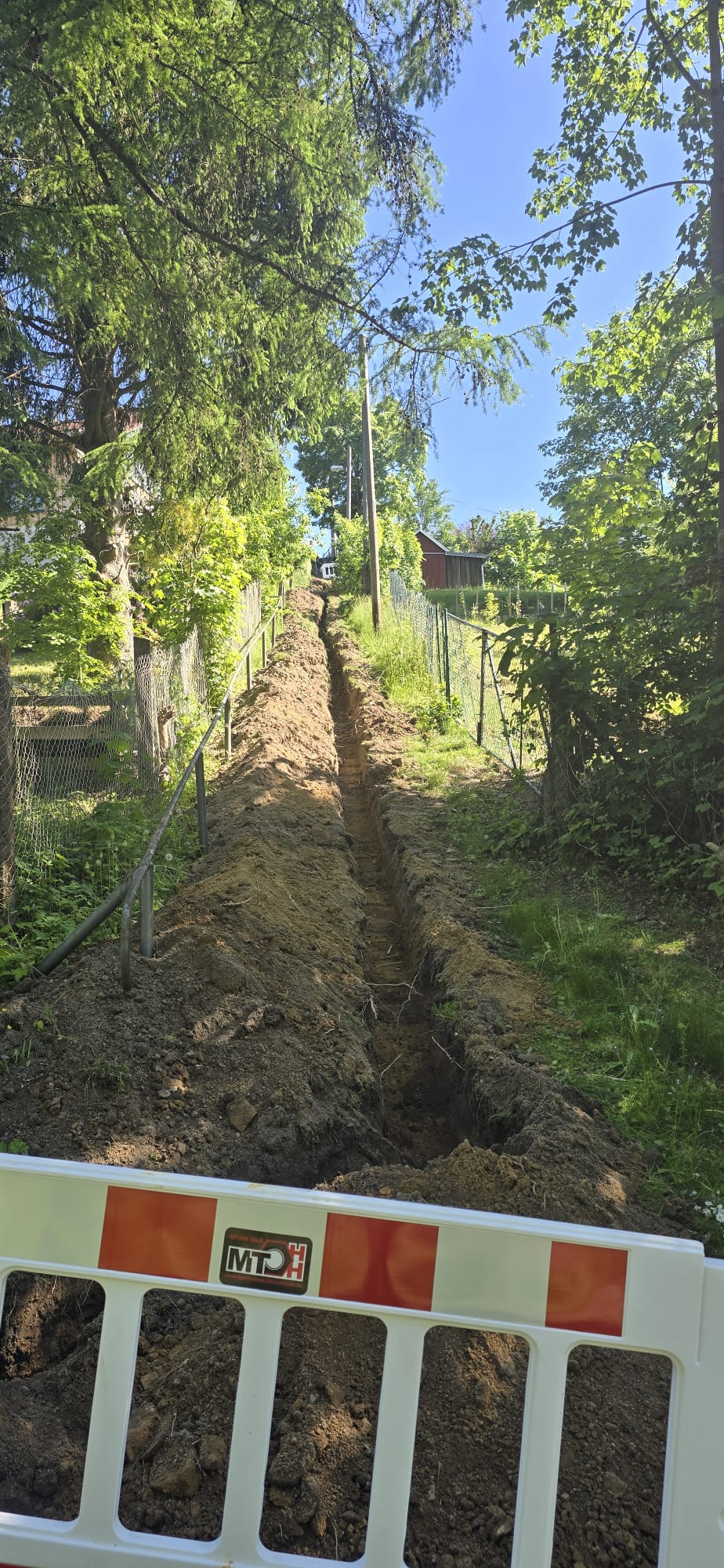 MTHH GbR - Kabeltragsystembau, Regalbau/Prüfung und Tiefbau - Zwischen zwei Zäunen wurde ein langer, schmaler Graben durch einen grasbewachsenen, von Bäumen gesäumten Hang gegraben. Im Vordergrund steht eine weiß-orange Bauabsperrung.