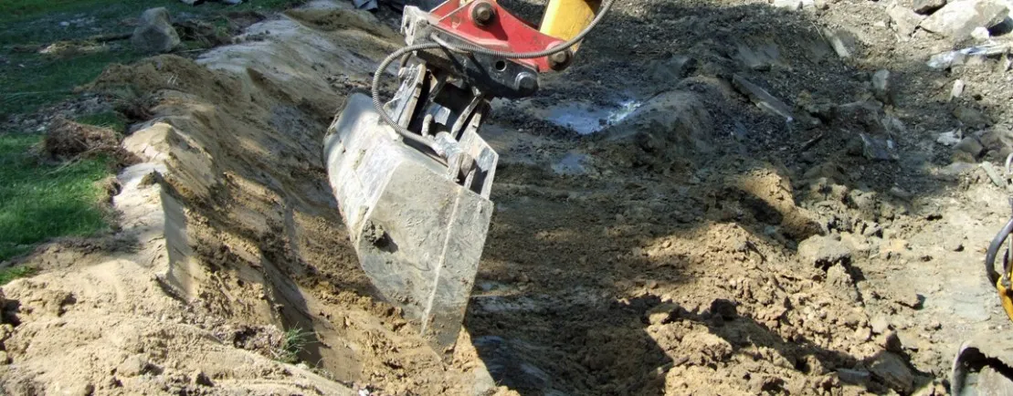 MTHH GbR - Kabeltragsystembau, Regalbau/Prüfung und Tiefbau - Ein Baggerlöffel gräbt einen Graben in die Erde auf einer Baustelle.