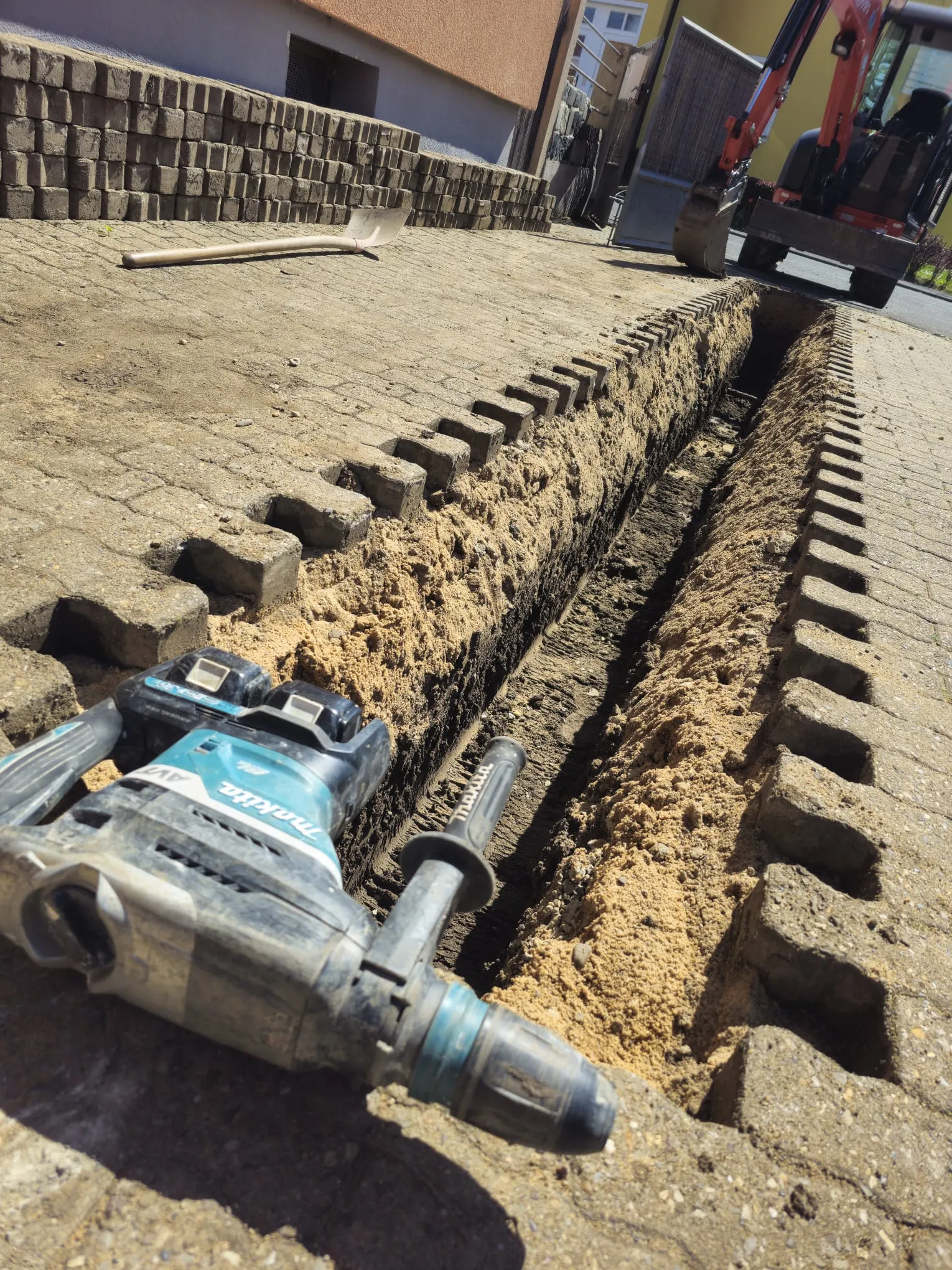 MTHH GbR - Kabeltragsystembau, Regalbau/Prüfung und Tiefbau - Auf einer gepflasterten Fläche neben einem schmalen, durch Ziegelsteine ​​gegrabenen Graben liegt ein Makita-Bohrhammer; im Hintergrund ist ein kleiner Bagger zu sehen.