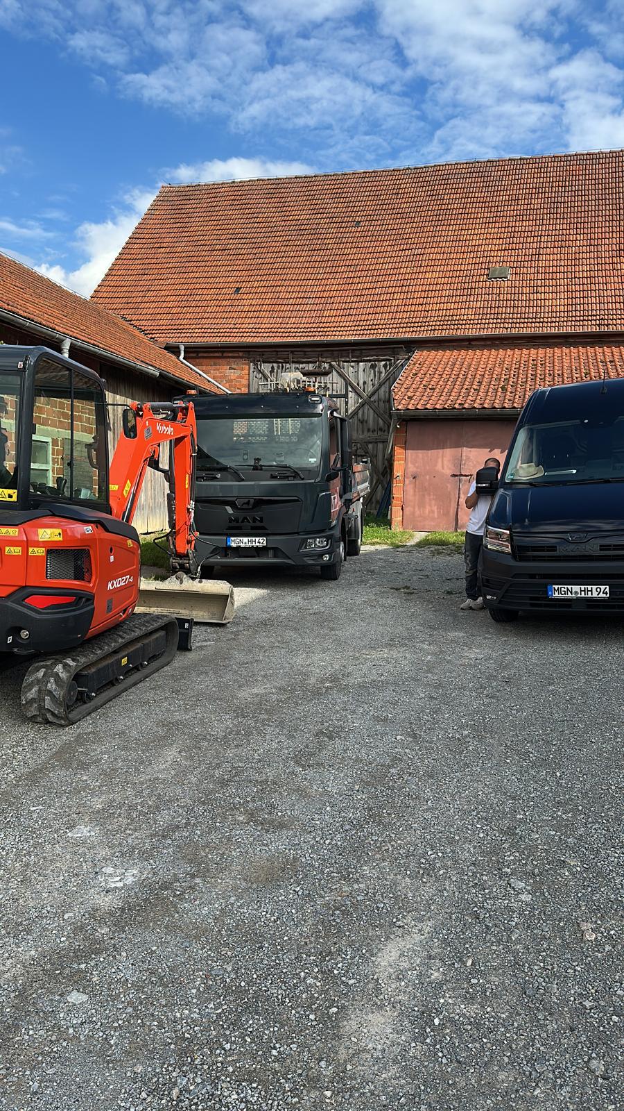 MTHH GbR - Kabeltragsystembau, Regalbau/Prüfung und Tiefbau - Ein kompakter roter Kubota-Bagger steht auf einem Schotterplatz neben einem schwarzen MAN-Lkw und einem dunklen Lieferwagen, die vor einer rustikalen Backsteinscheune mit einem großen orangefarbenen Ziegeldach positioniert sind.